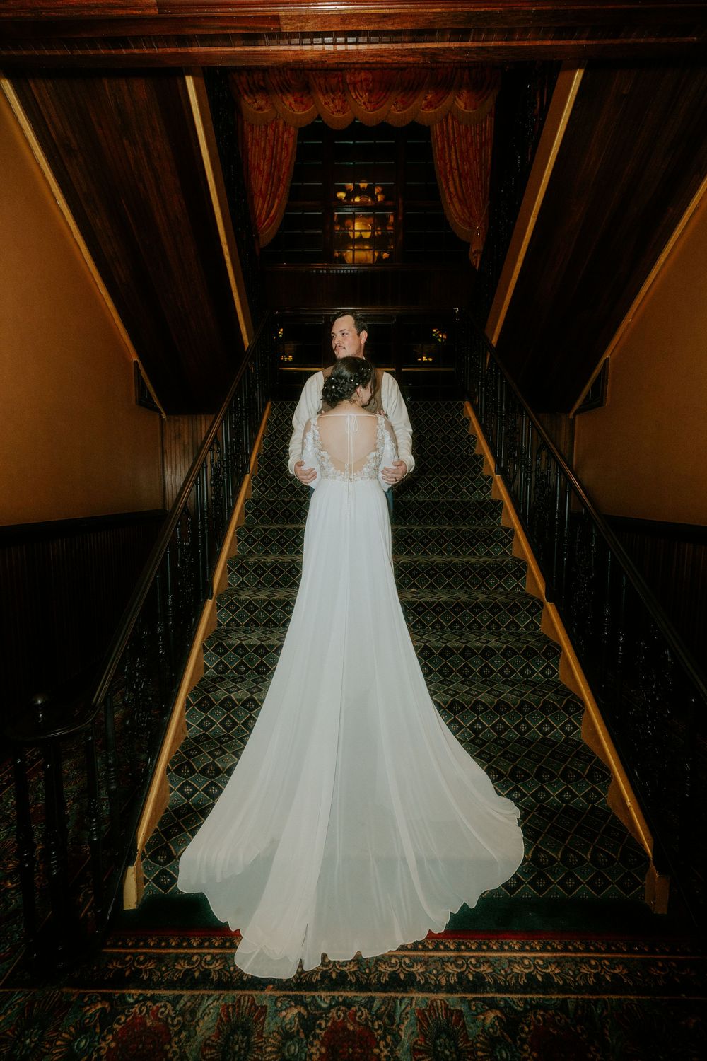 Bride in elegant white gown with lace back and train stands on ornate staircase in grand venue.