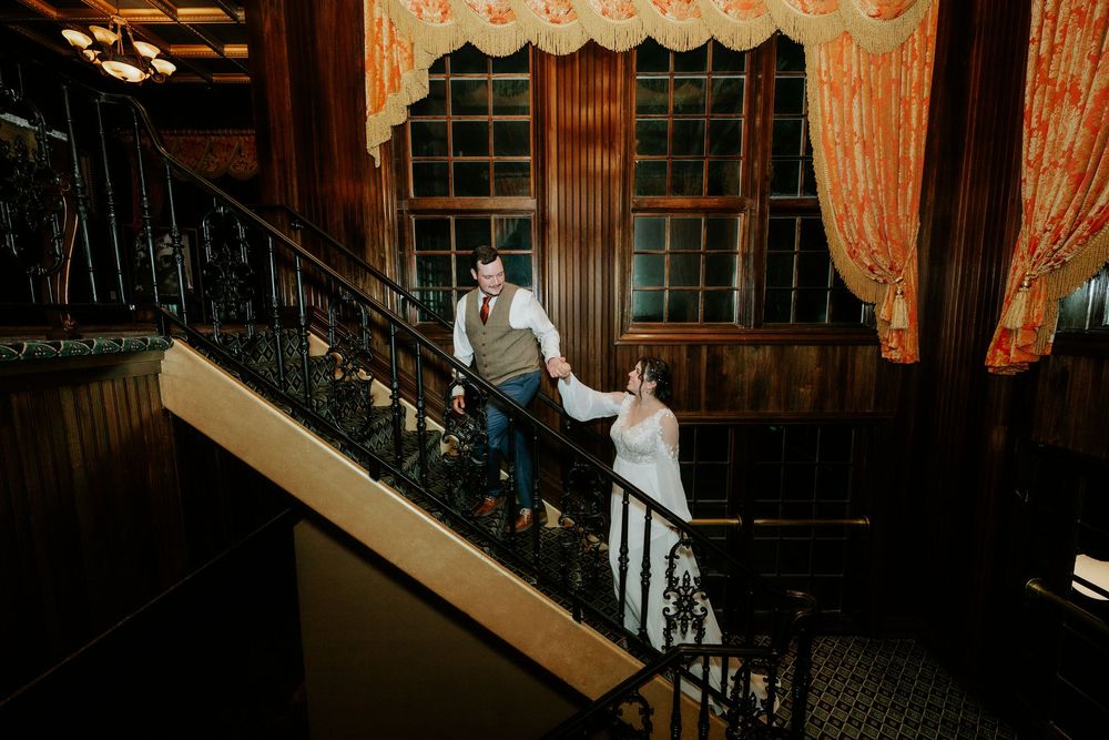 Couple in elegant attire poses on ornate staircase with wrought iron railings in a warmly lit historic venue.