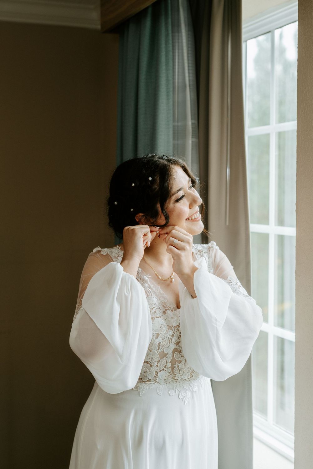 A smiling bride in a white lace gown with billowy sleeves puts on earrings by a sunlit window.