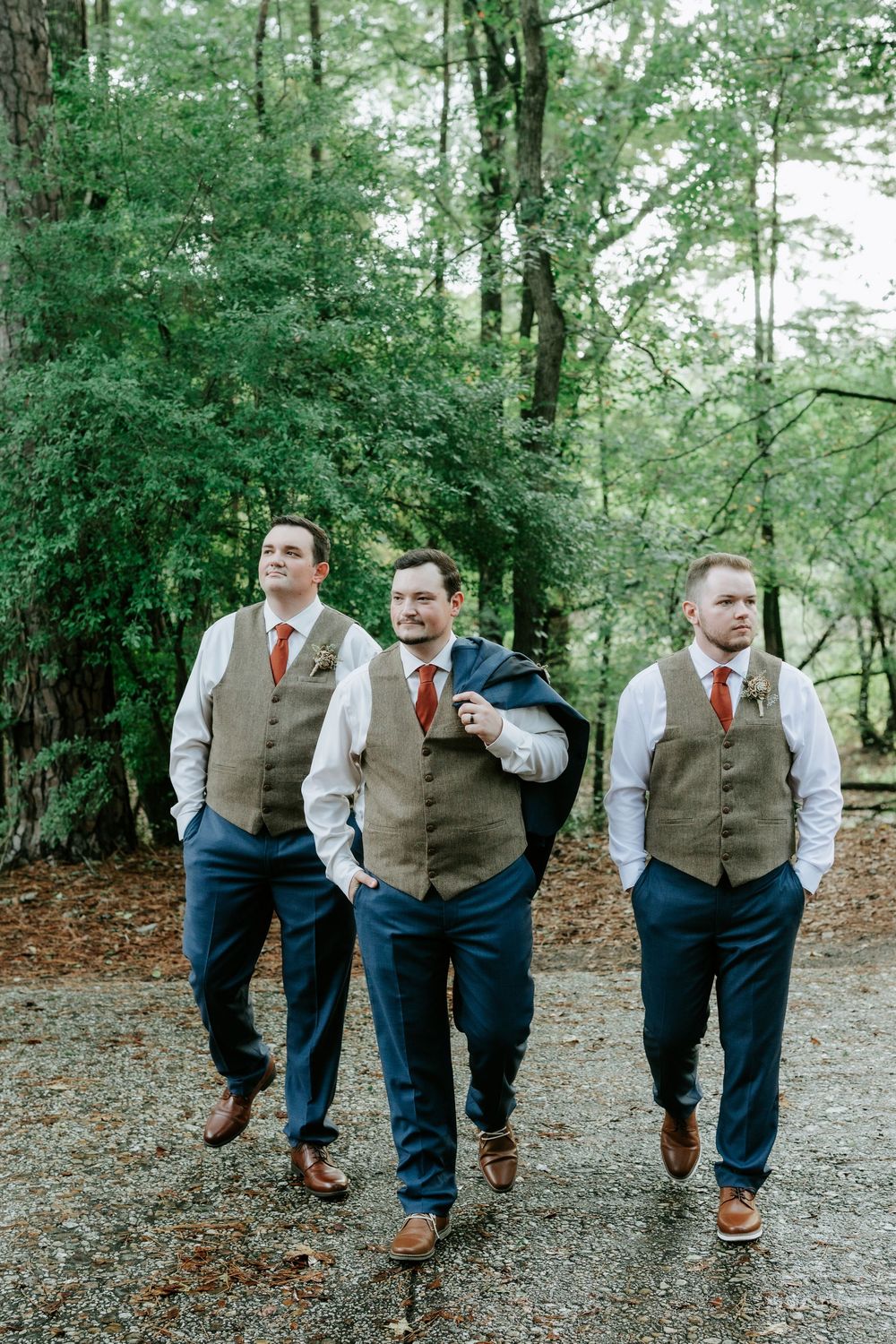 Three groomsmen in tan vests, navy pants, and red ties walk together outdoors in a wooded setting.