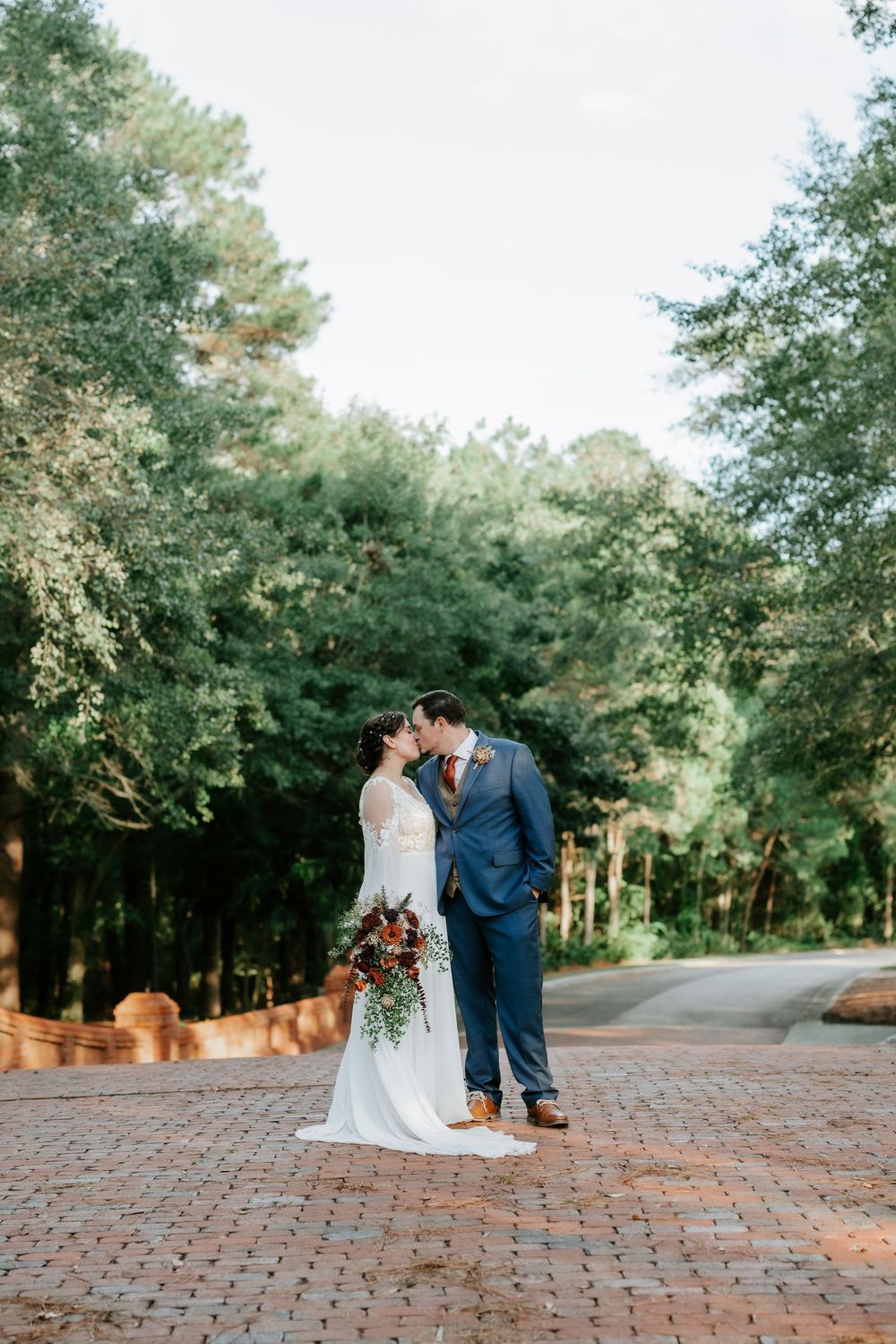 Bride and groom share a kiss on a brick path surrounded by lush green trees on their wedding day.