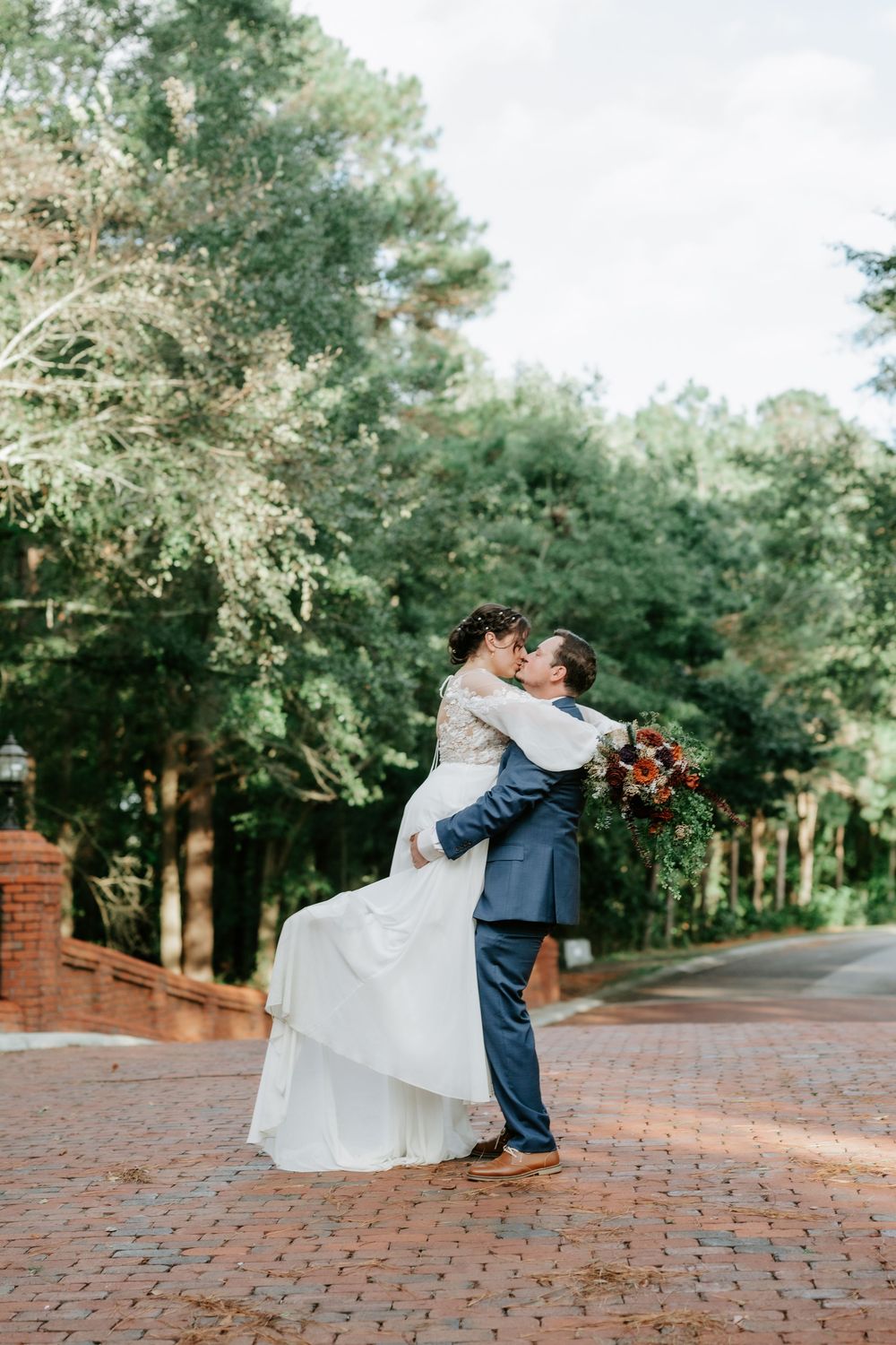 Groom lifts bride in white gown on brick path, sharing a kiss surrounded by lush green trees at outdoor wedding.
