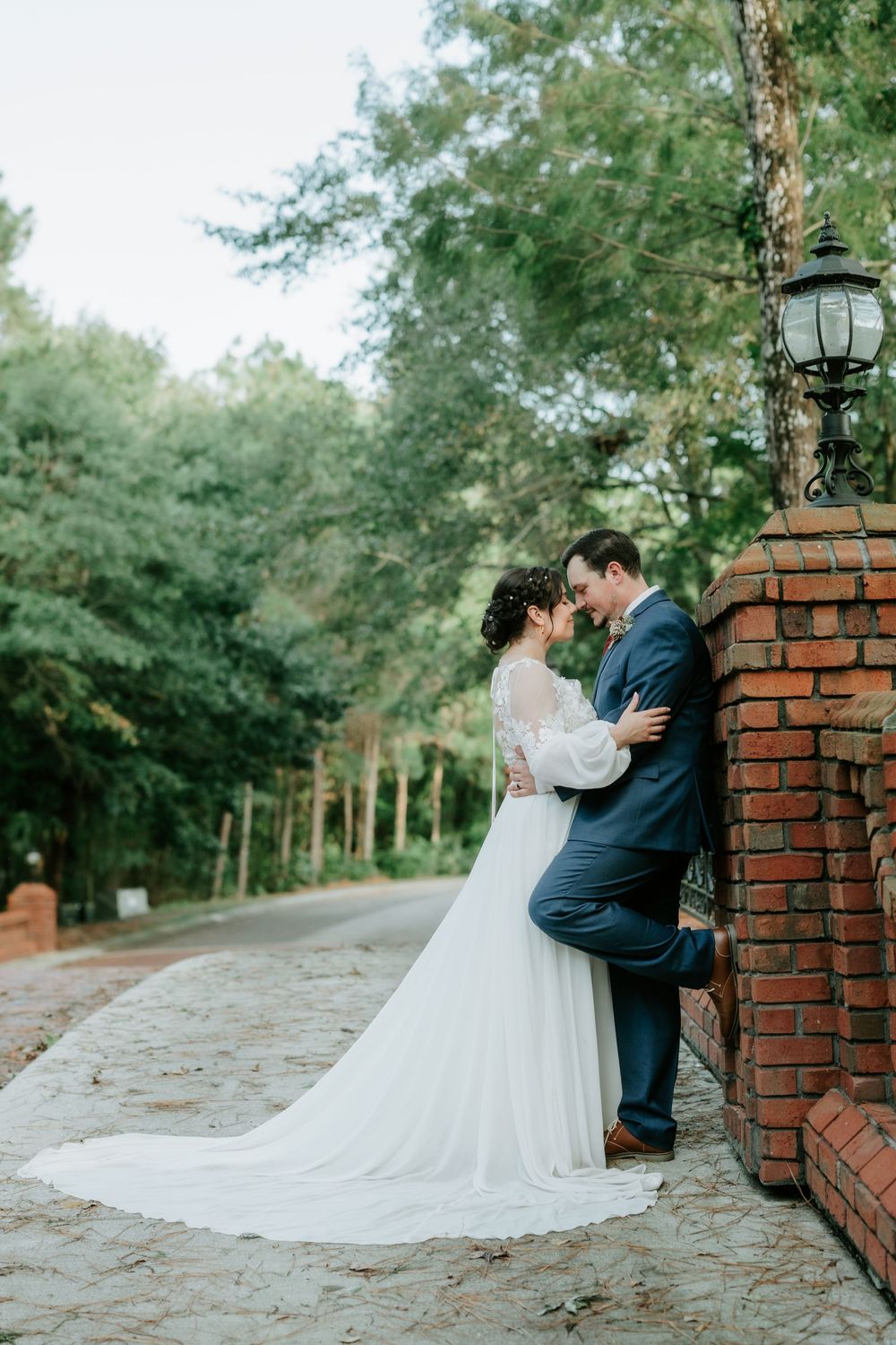 Bride and groom share romantic moment by brick pillar surrounded by lush green trees outdoors.