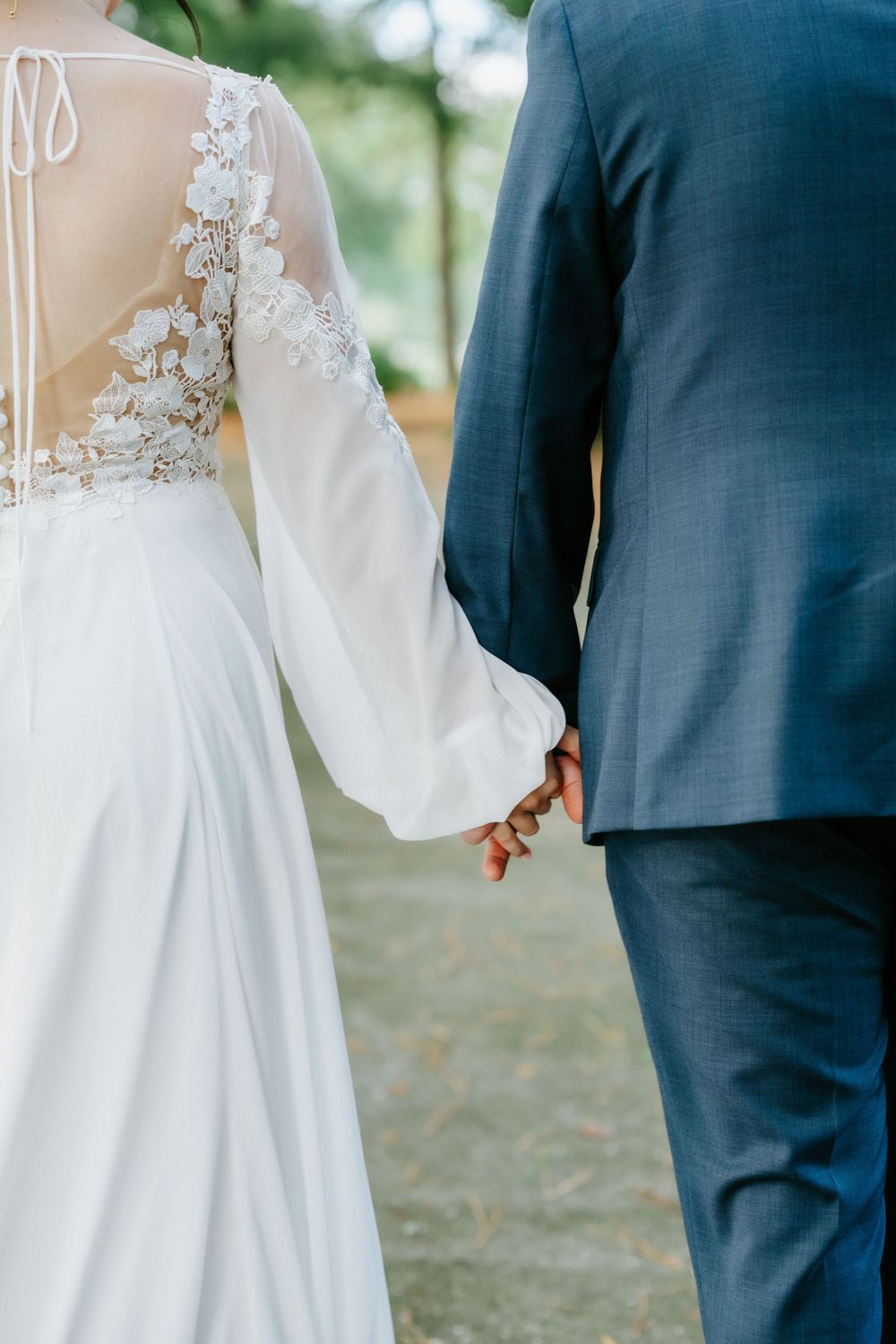 Bride and groom holding hands outdoors, bride wearing elegant lace-back white wedding gown, groom in blue suit.