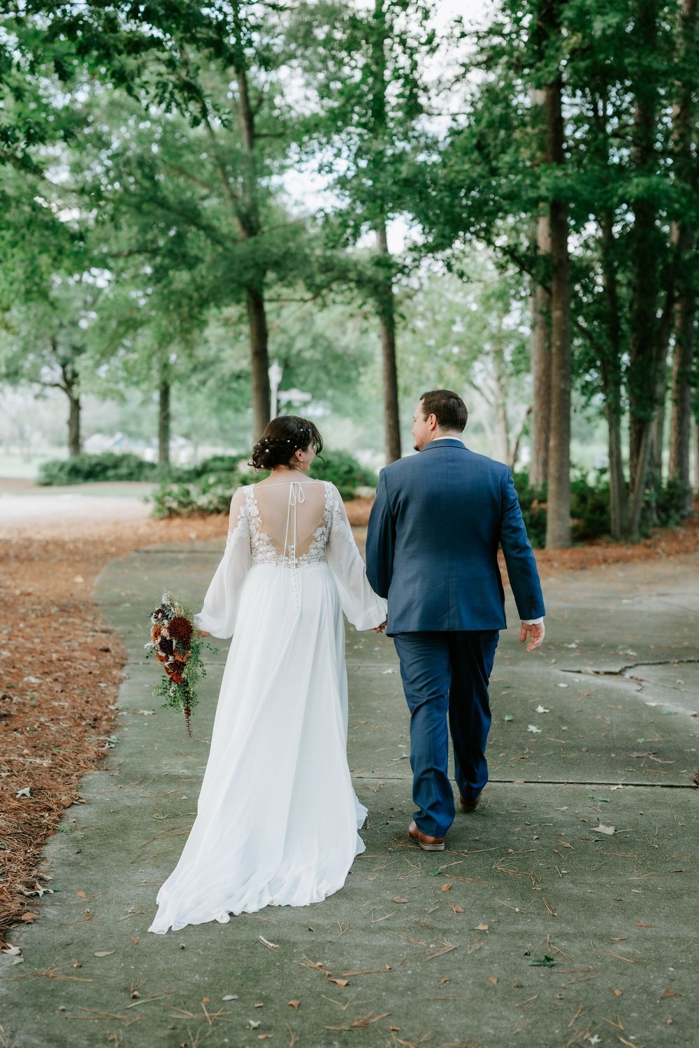 A bride and groom walk hand in hand down a tree-lined path, she in a white gown holding a bouquet, he in a navy suit.