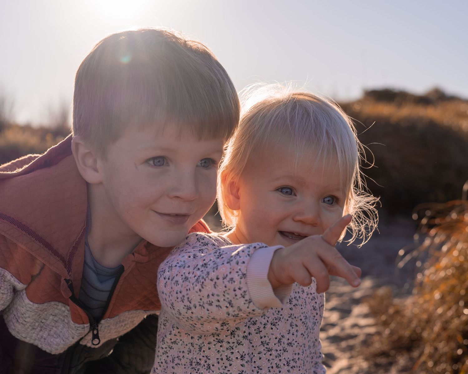 Two young siblings smiling outdoors in golden sunlight, older child hugging a pointing toddler near sandy dunes.