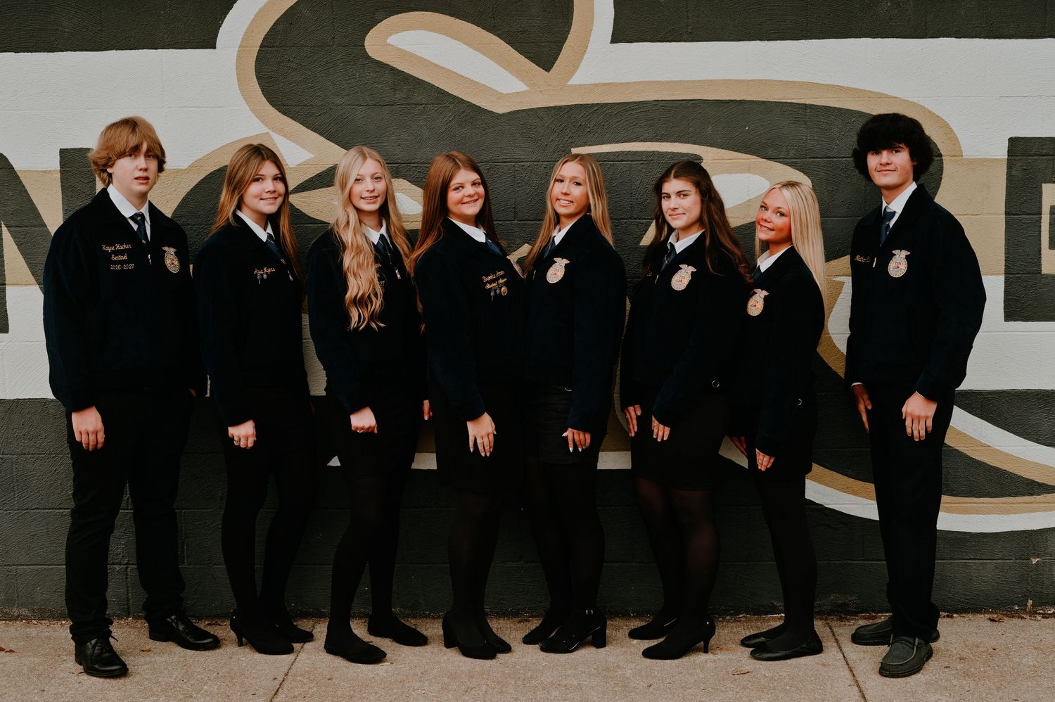 Eight FFA students in matching navy blue jackets pose together outdoors in front of a painted wall.