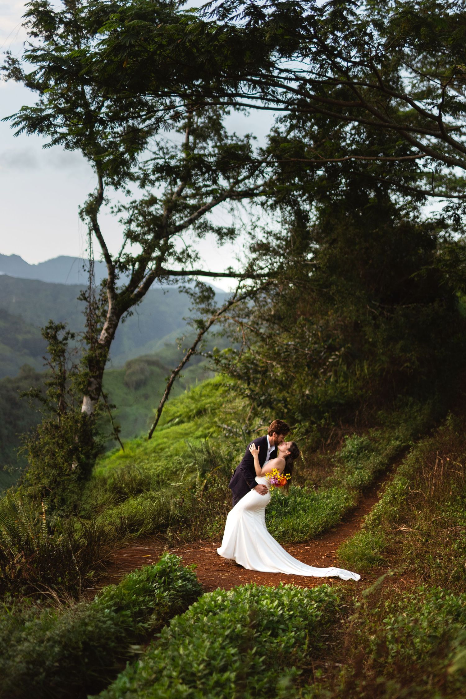 Bride in a white gown and groom in a dark suit share a dipped kiss on a winding dirt path surrounded by green mountains and trees.