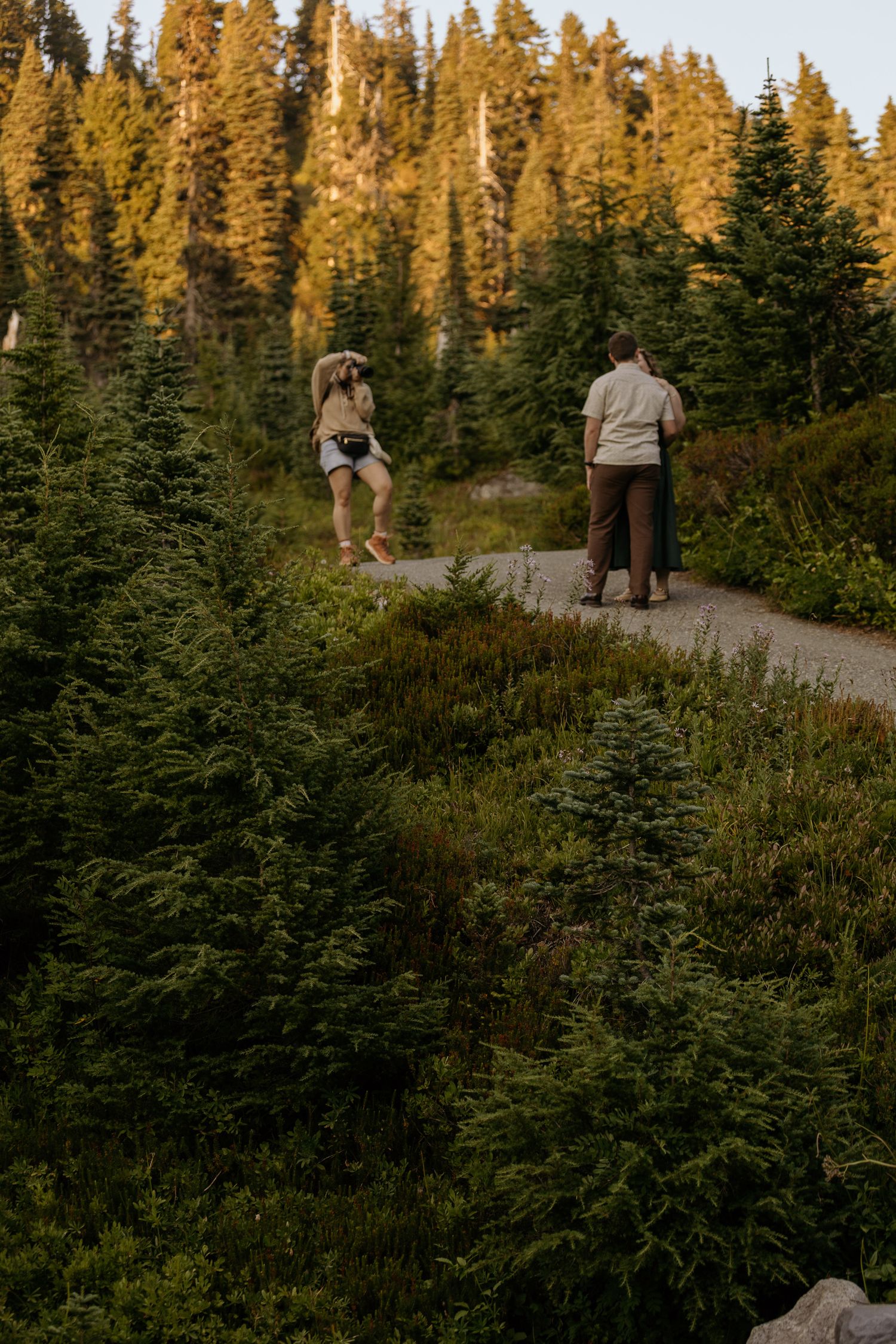 Photographer crouching to capture a couple standing close together on a gravel mountain trail surrounded by evergreen trees and wild shrubs.