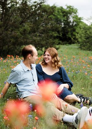 Happy couple laughs together while sitting in a meadow of blooming wildflowers, with blurred orange blossoms in the foreground.