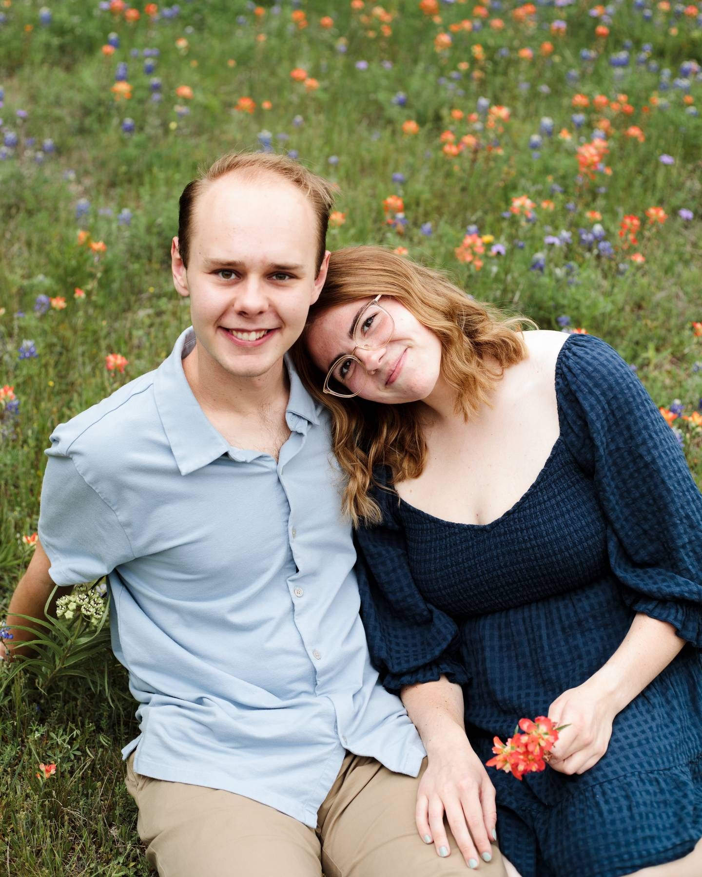 Smiling couple sits close together in a field of orange and purple wildflowers during an outdoor engagement photo session.
