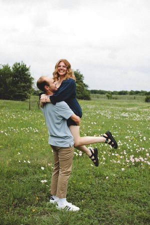 Man lifts his smiling fiancée off the ground in a joyful embrace in a green field dotted with white wildflowers.