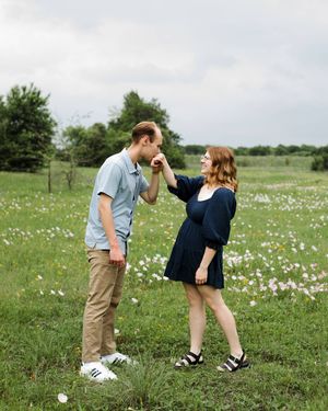 Man gently kisses his partner's hand as they stand in a grassy wildflower field under an overcast sky in an engagement photo.