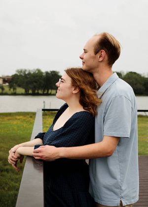 Engaged couple embraces at a lakeside railing, the woman leaning back against her partner as they gaze out over the water.