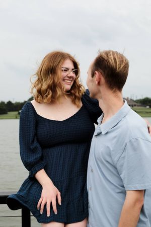A smiling woman in a navy dress sits on a railing beside a man in a light blue shirt, with a lake behind them.