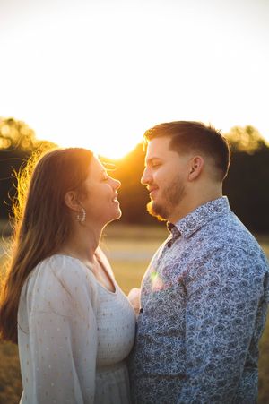 A couple stands face to face smiling at each other in warm golden sunset light during an outdoor engagement photo shoot.