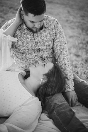 A black and white photo of a woman resting her head on a man's lap while he gently touches her hair outdoors.