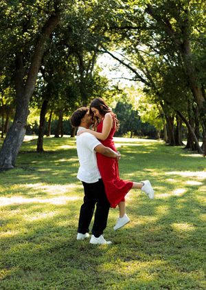 A man lifts a woman in a red dress into an embrace beneath tall shady trees in a sunlit park.