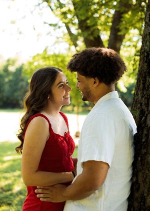 A smiling woman in a red dress leans into a man in a white shirt as they stand together beside a tree outdoors.