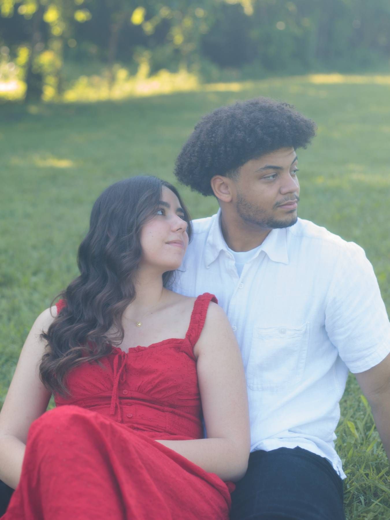 A couple in a red dress and white shirt sit close together on the grass, gazing off in a softly lit park.