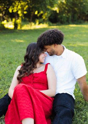 A man touches foreheads with a smiling woman in a red dress as they sit together on a grassy lawn.