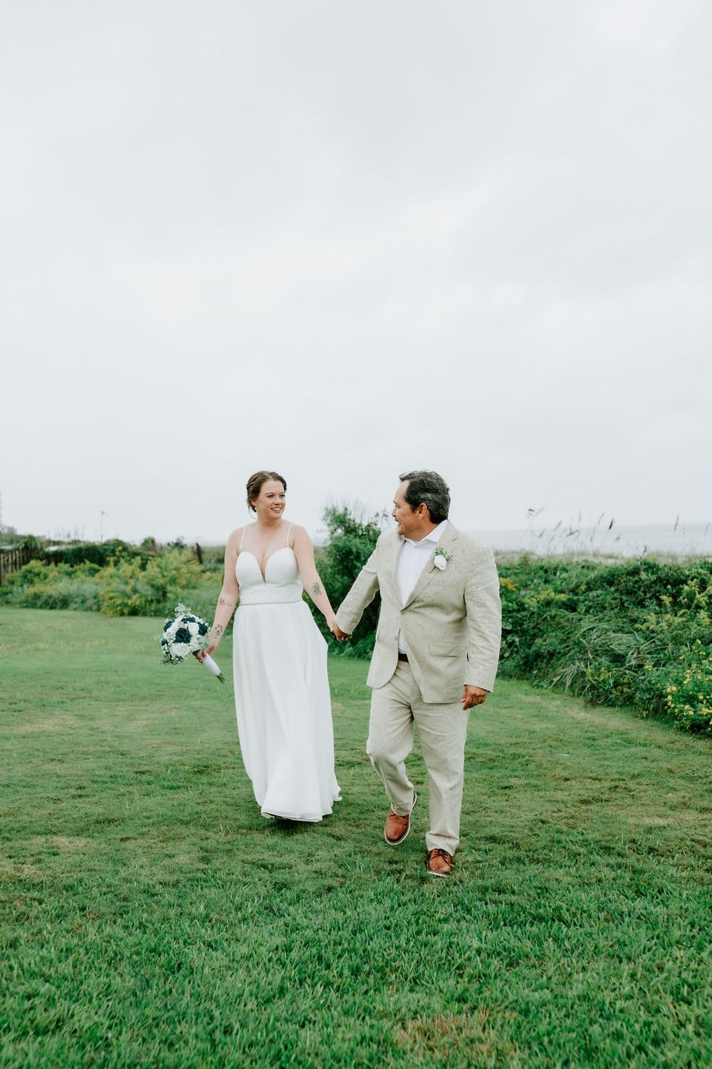Bride in a white gown and groom in a tan suit hold hands while walking across a green lawn at a seaside wedding.