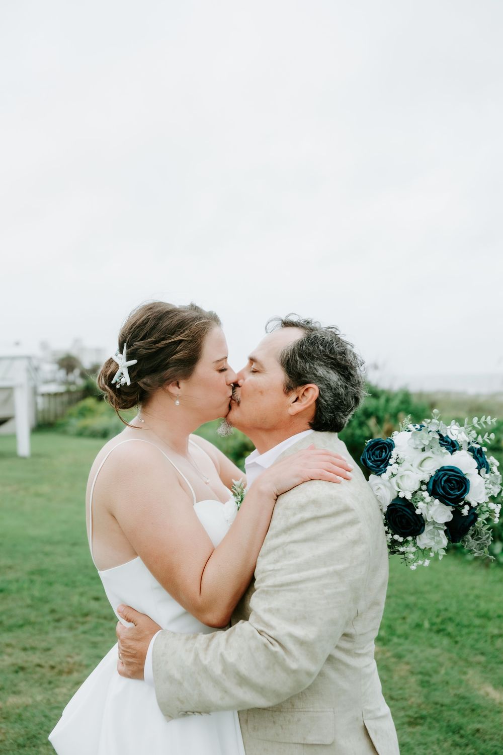 Bride in a white dress and groom in a tan blazer share a kiss on a grassy lawn, holding a navy and white floral bouquet.