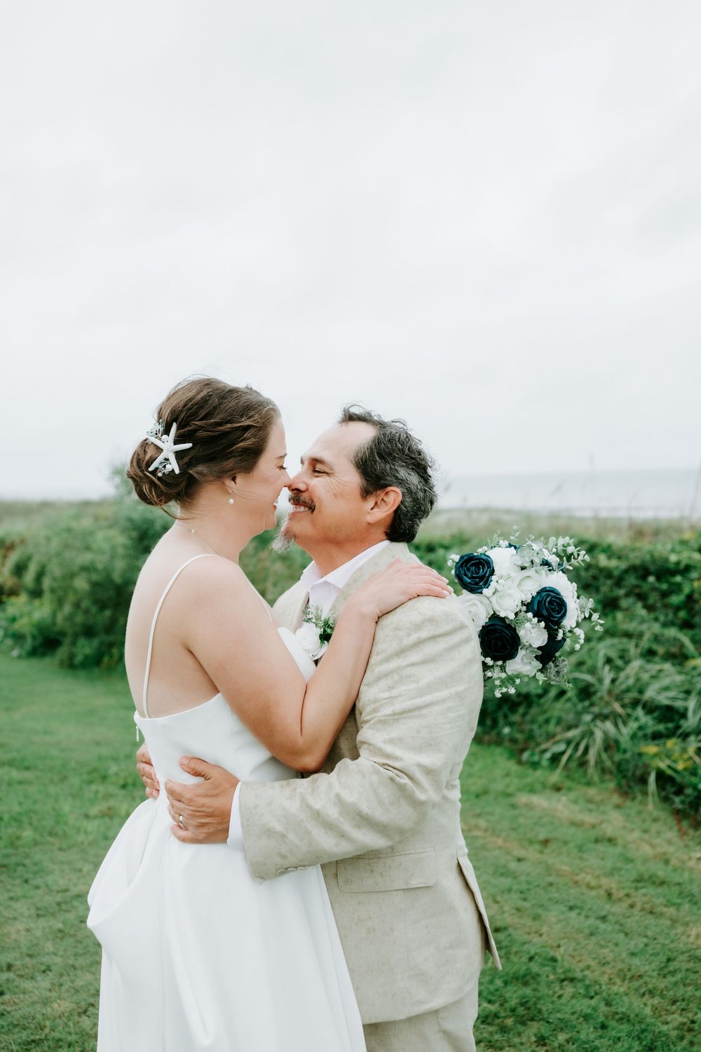 Bride in a white dress with a starfish hairpin embraces her groom in a tan suit on a grassy coastal lawn, holding a navy and white bouquet.