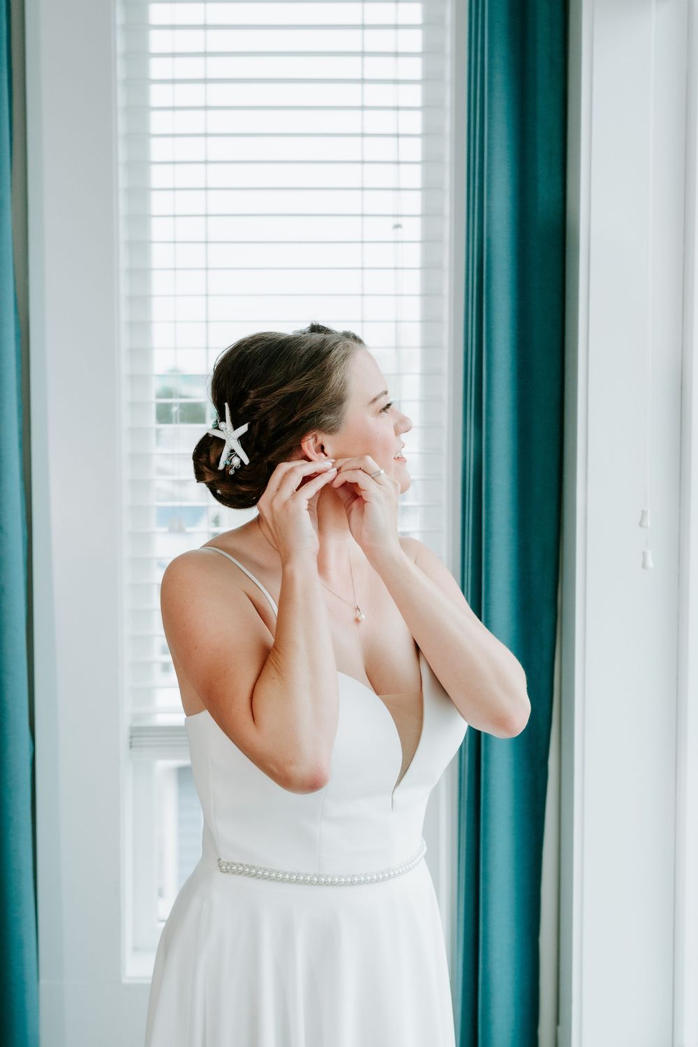 Bride in a white gown with a starfish hair clip adjusts her earring beside a sunlit window with teal curtains.