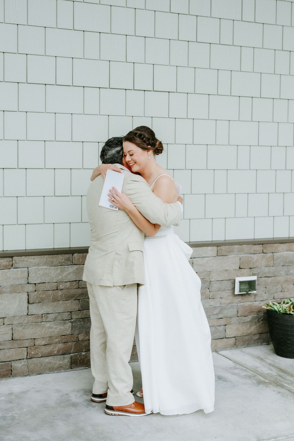 A bride in a white gown and a groom in a beige linen suit share a warm embrace against a white brick and stone wall.