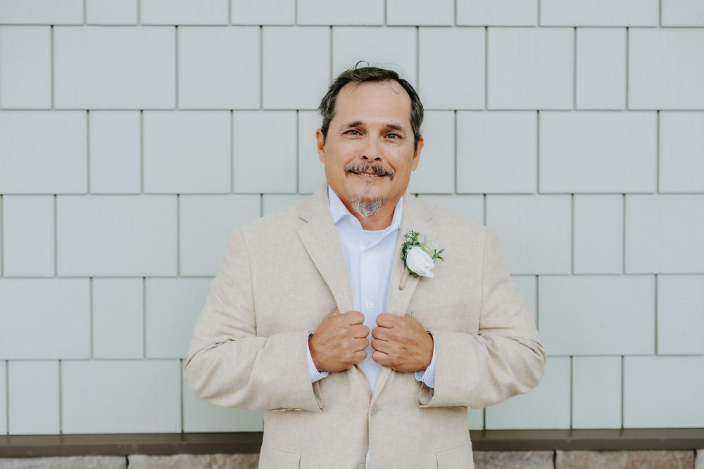 Smiling groom in a tan linen suit with a white floral boutonniere stands adjusting his jacket in front of a white shiplap wall.