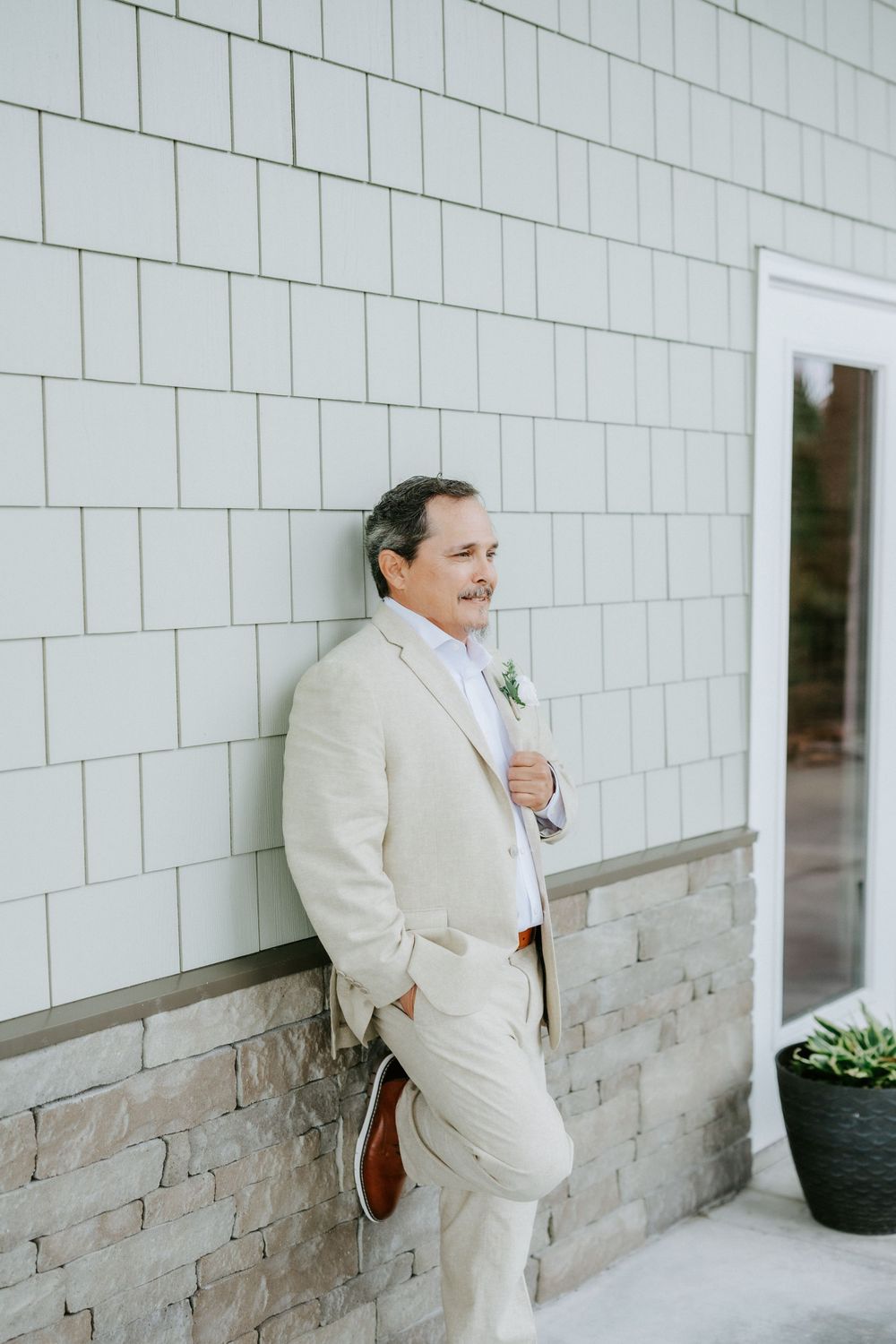 Smiling man in a beige linen suit with a boutonniere leans against a gray shingled wall above a stone base, one hand in his pocket.