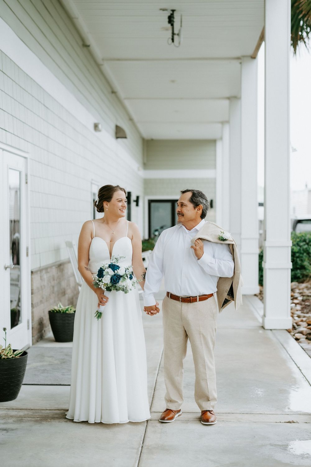 Smiling bride in a white gown holding a blue bouquet walks hand in hand with her groom on a covered porch at a coastal wedding.