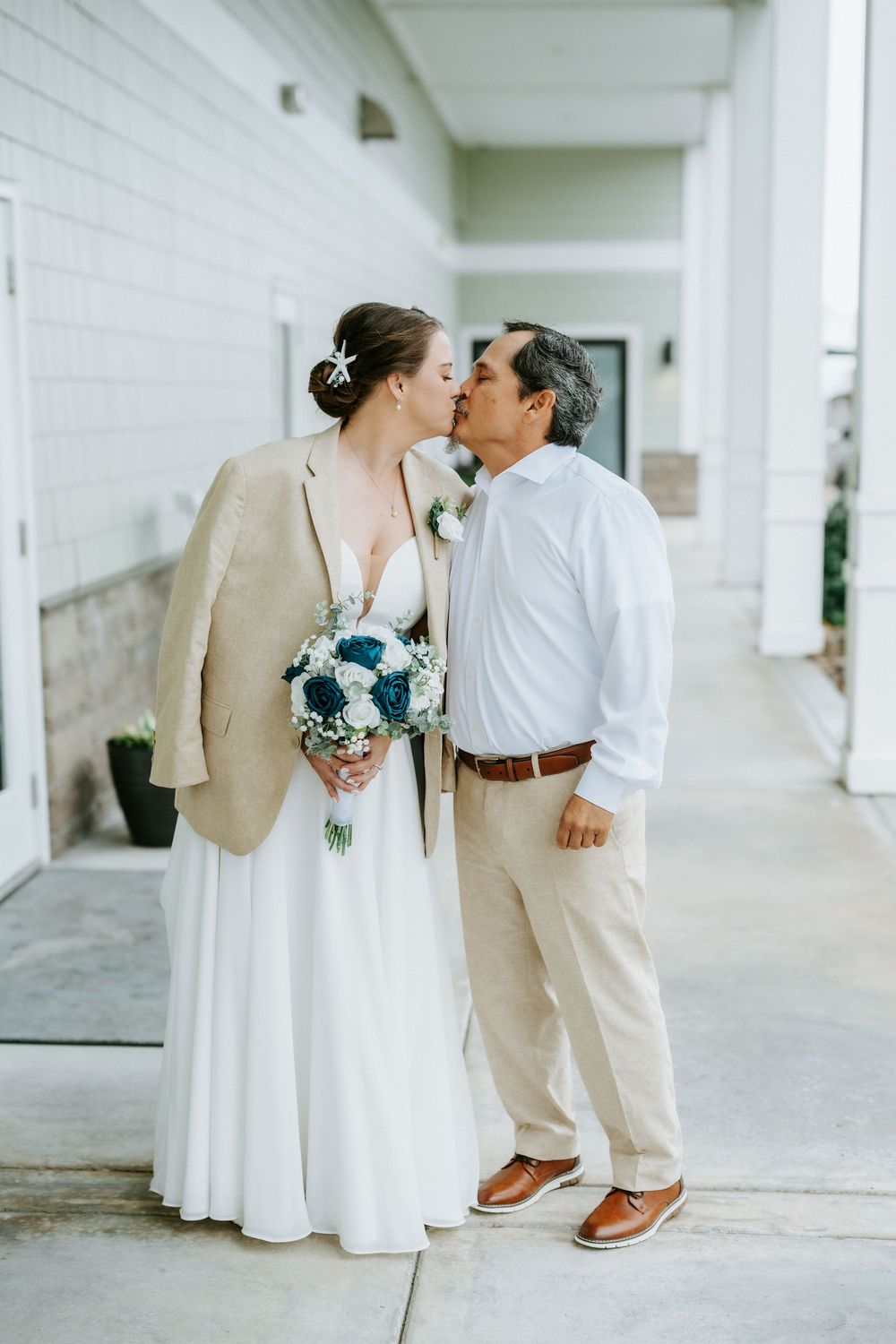 Bride in a white dress and tan blazer holding a blue and white bouquet kisses her groom in khakis on a covered wedding venue porch.