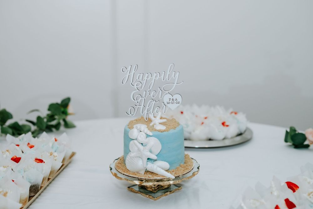 Small blue celebration cake with white flowers and a Happy Anniversary topper on a white table beside frosted cupcakes and green leaves.