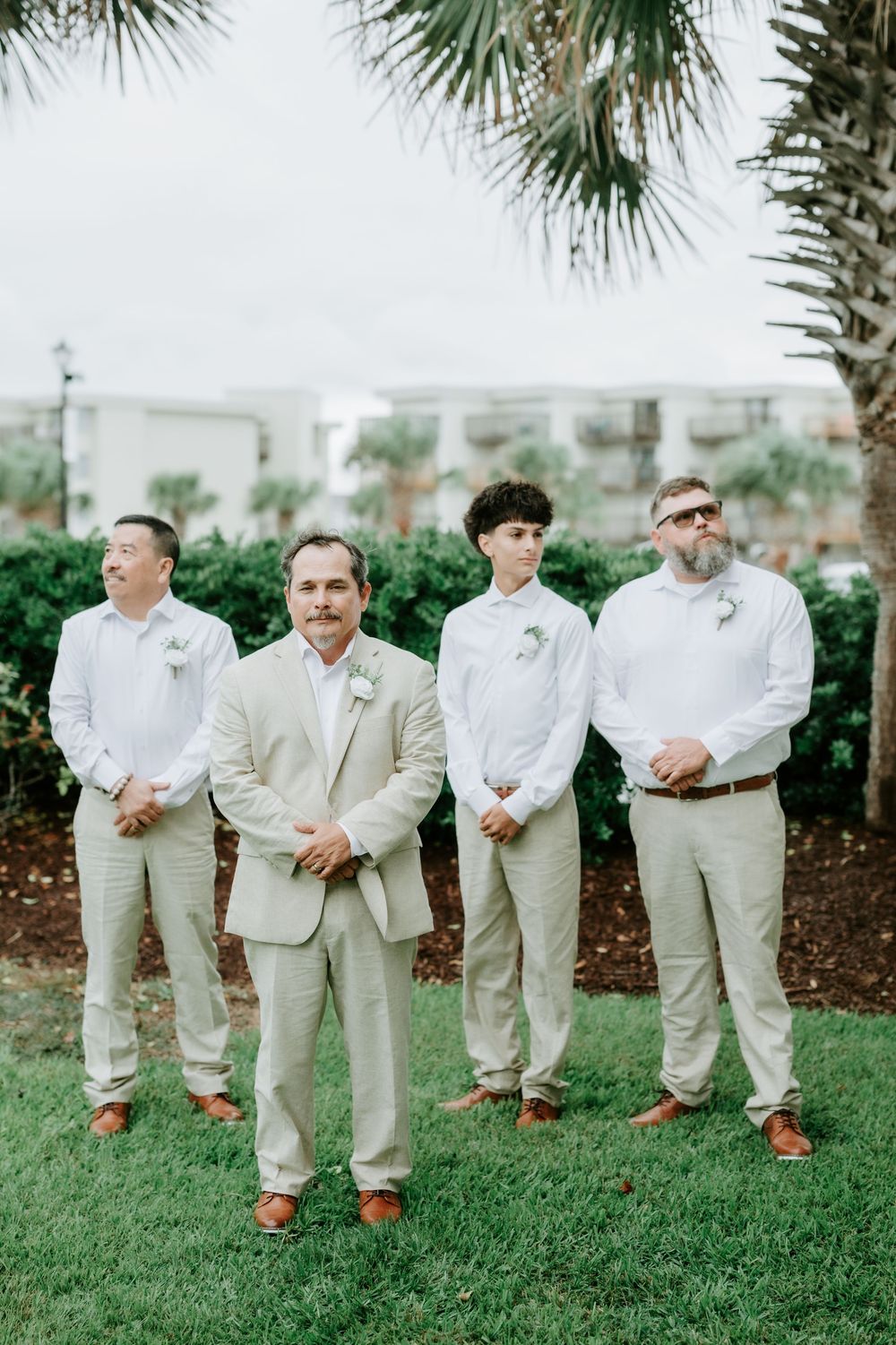 Four groomsmen in tan suits and white shirts with boutonnieres stand on a green lawn beneath palm trees at a beach wedding.