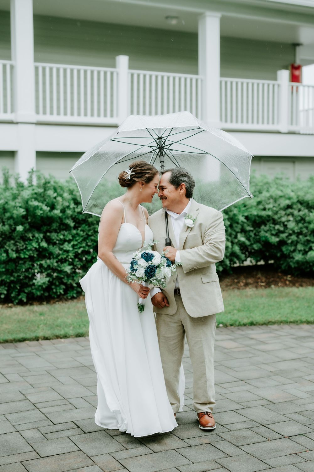 A bride in a white gown and a groom in a tan suit touch foreheads under a clear umbrella, holding a blue bouquet on a brick patio.