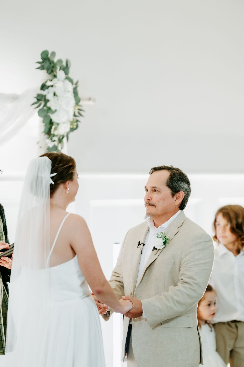Bride in a white gown and veil holds hands with a groom in a tan suit during a wedding ceremony as children look on.