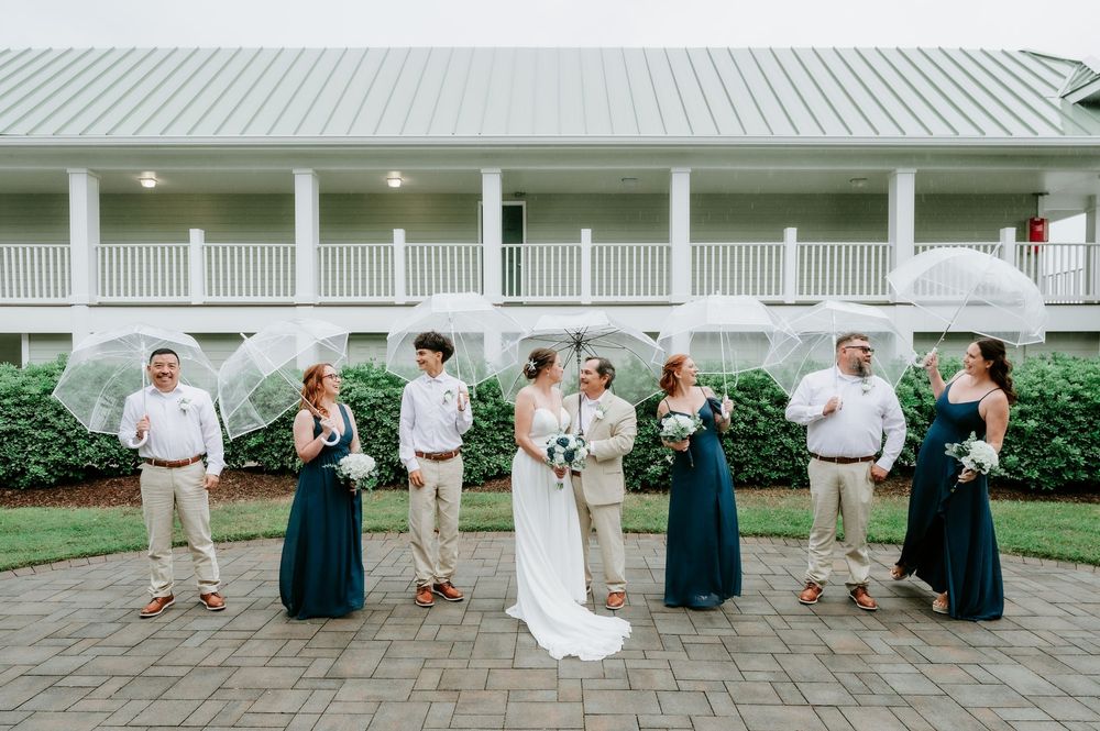 Bride and groom stand with their wedding party holding clear umbrellas on a brick patio in front of a white porch building.
