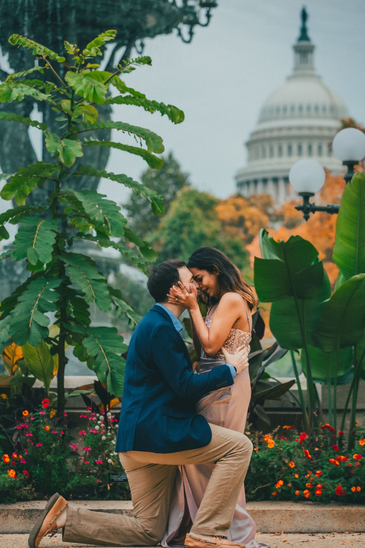 Man in a navy blazer kneels on one knee proposing to a smiling woman near tropical plants with the US Capitol dome in the background.