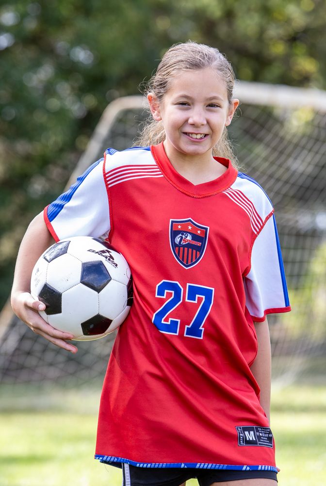 Smiling young soccer player in a red number 27 jersey holds a soccer ball in front of a goal net on a sunny grass field.