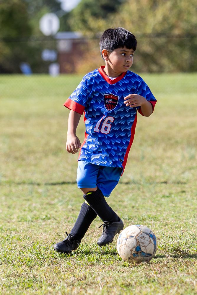 Young boy in a blue number 16 soccer jersey and black socks dribbles a soccer ball across a grassy field on a sunny day.