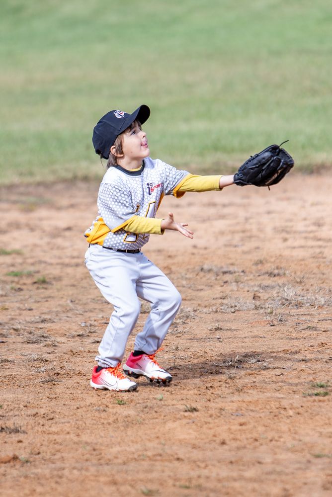 Young youth baseball player in a gray and yellow uniform and cap reaches up with a black glove to catch a fly ball on a dirt infield.