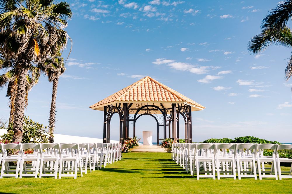 Oceanfront wedding gazebo with a tile roof, framed by palm trees and rows of white chairs on a green lawn under a blue sky.