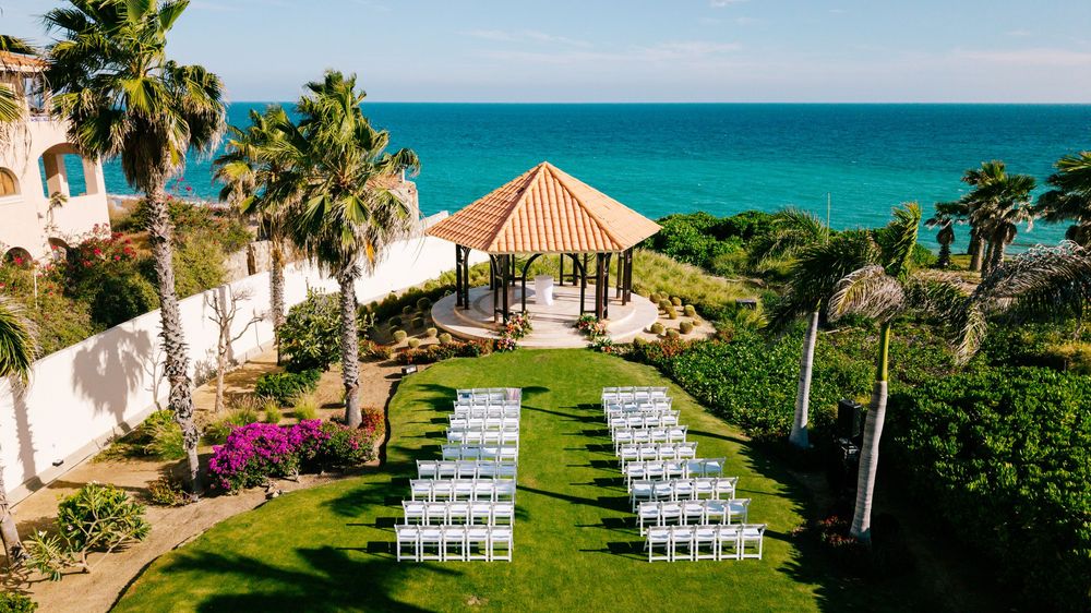 Oceanfront wedding ceremony setup with white chairs on a green lawn facing a wooden gazebo, surrounded by palm trees and flowers.