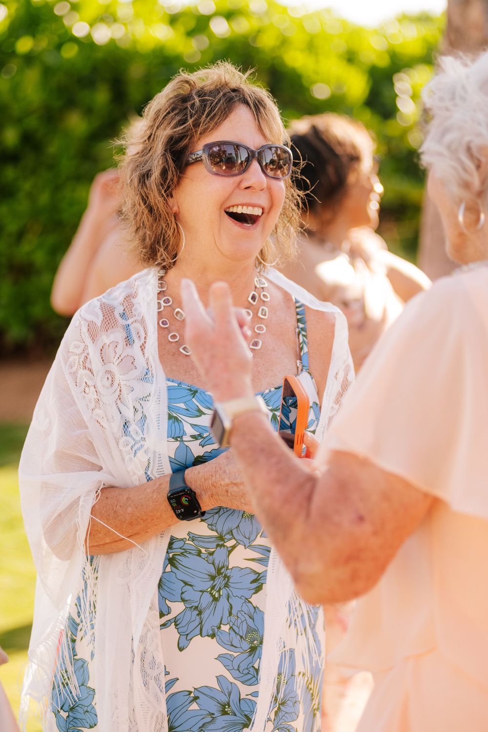 Smiling woman in sunglasses, a blue floral dress and white lace shawl laughs while chatting with a friend at an outdoor garden gathering.