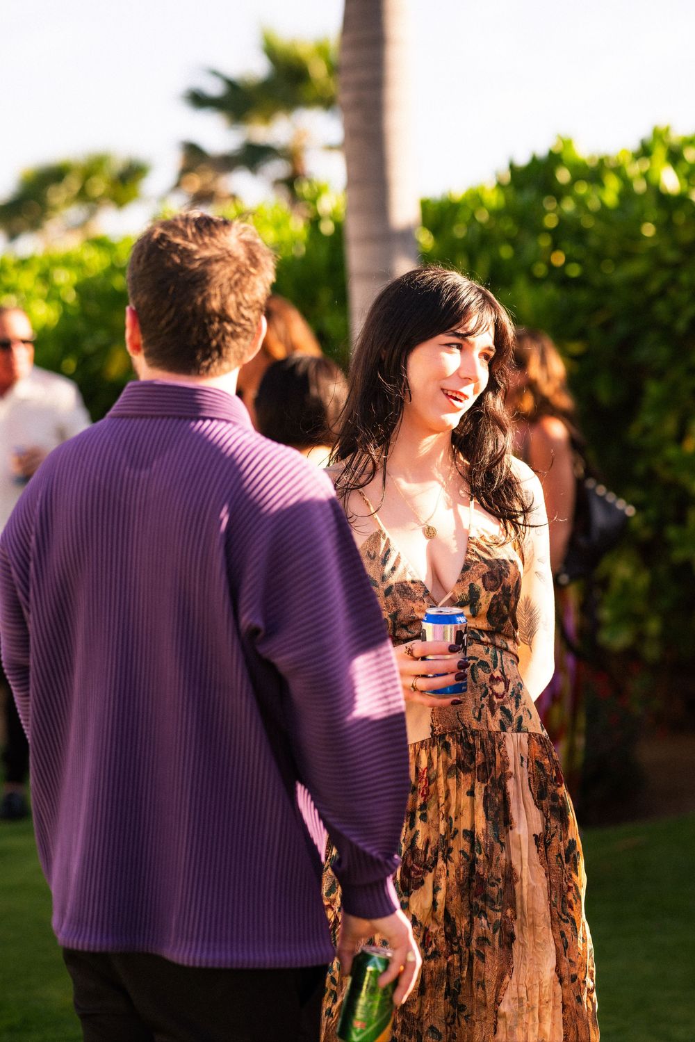 A woman in a floral dress holds a drink while chatting with a man in a purple sweater at an outdoor garden party with palm trees.