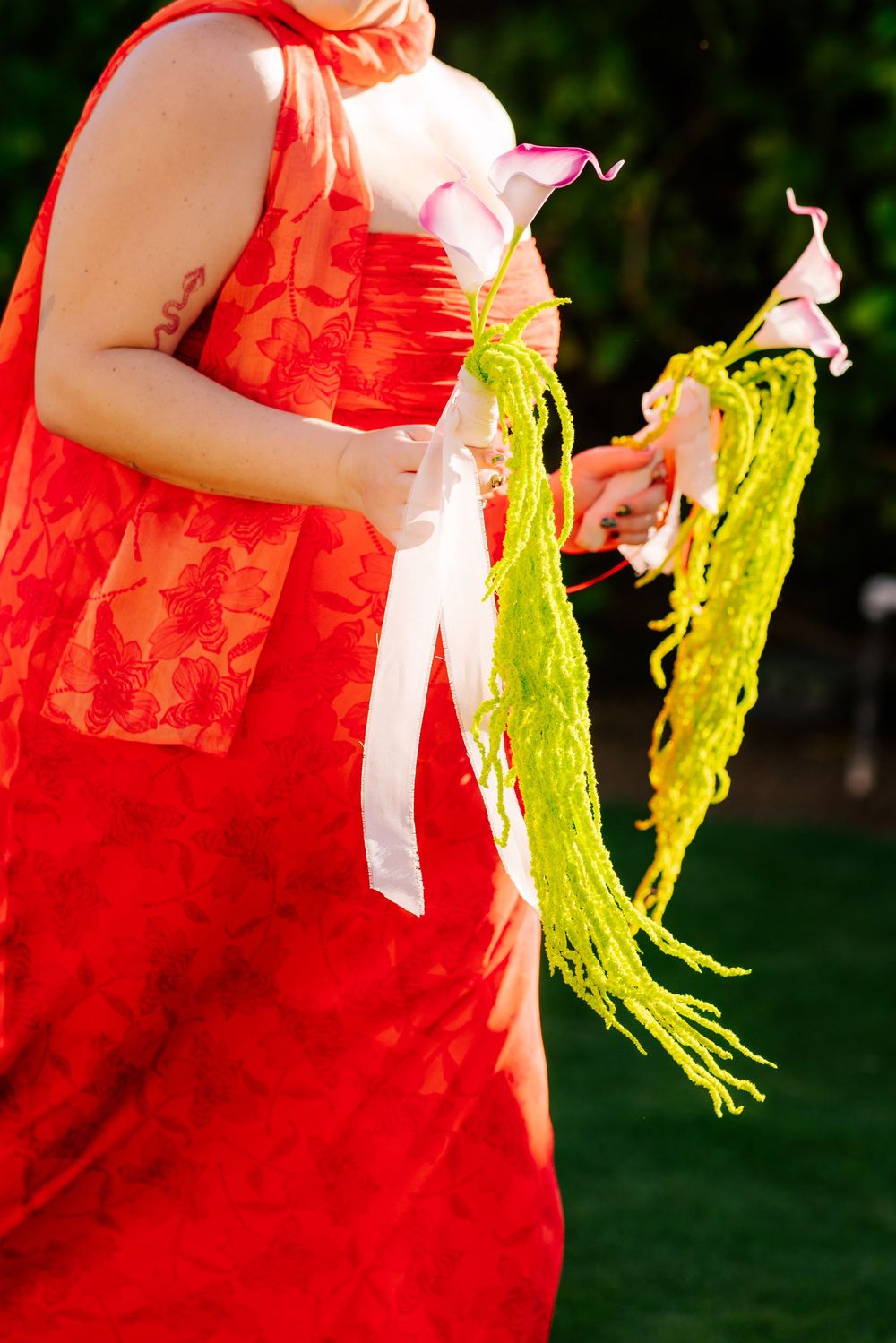 Bridesmaid in a red floral dress holding a bouquet of pink calla lilies with trailing green amaranthus and white ribbon on a lawn.