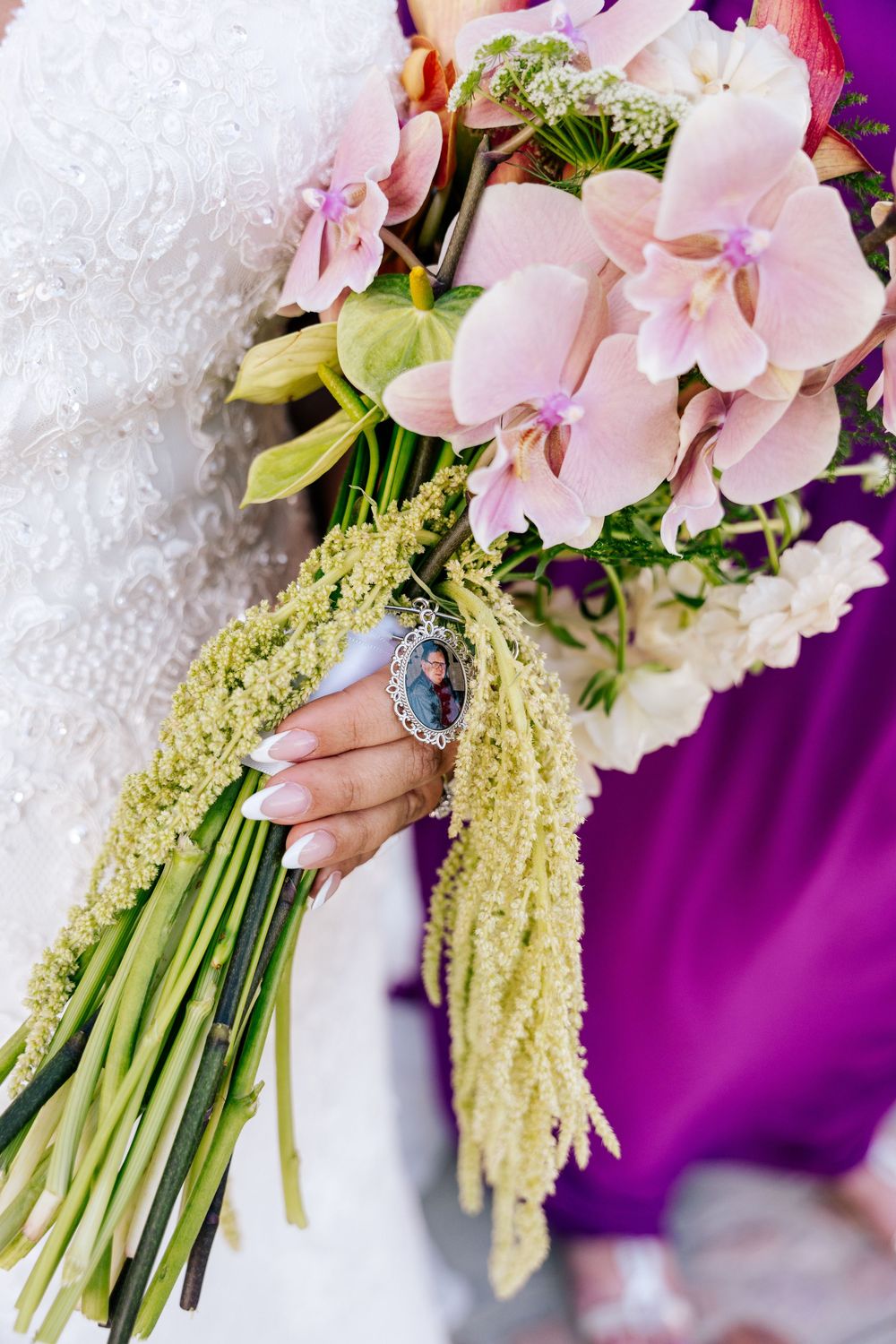 Bride in white lace gown holds a pink orchid bridal bouquet with green amaranthus and a photo memorial charm, beside a bridesmaid in purple.