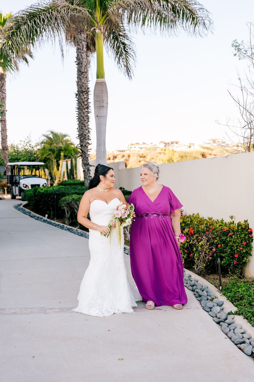 Bride in a white gown holding a bouquet walks arm in arm with a woman in a purple dress along a palm-lined path at a resort wedding.